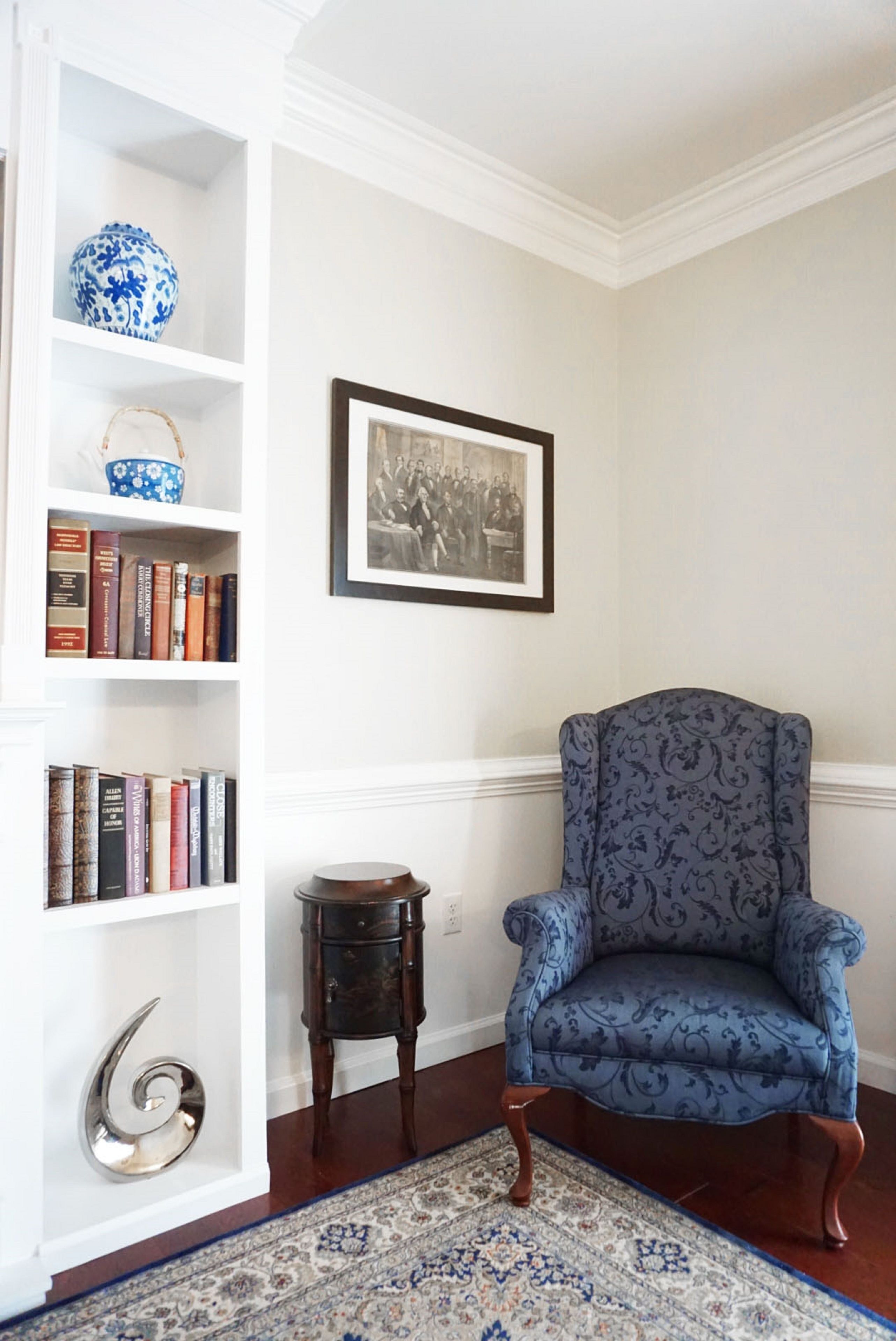 A room with a blue patterned armchair, a bookshelf with books and decorative items, a framed black and white photograph, and a patterned rug.