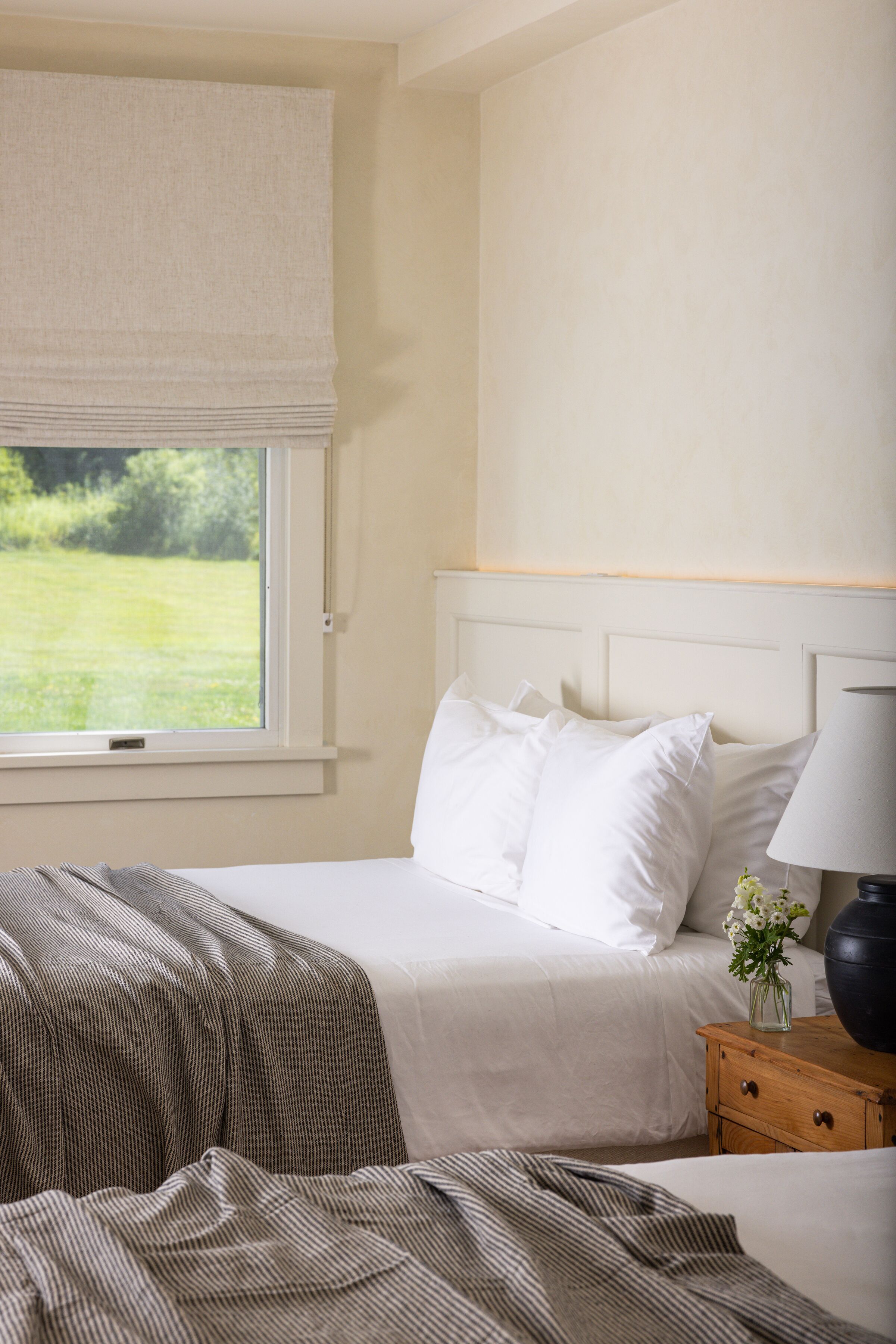 Una cama bien tendida con ropa blanca, una mesita de noche con lámpara, y vista a un paisaje verde a través de una ventana con persiana blanca.