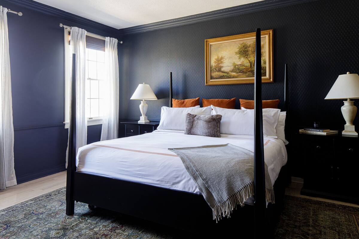 A neatly made bed with white linens and a gray throw blanket, a painting on the wall, and a window with curtains.