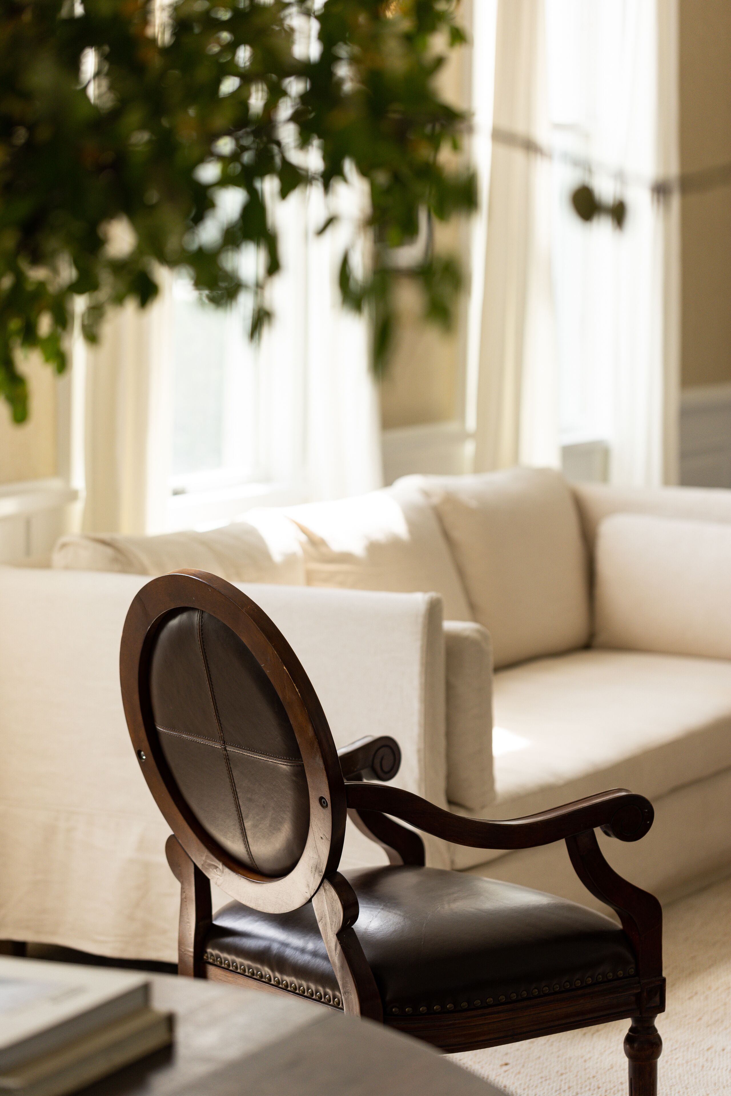 A room with a dark wooden chair, a white sofa, and a large window with curtains.