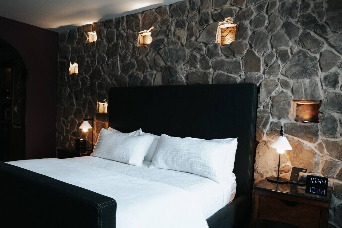 A bed with white linens, a stone wall, and wall-mounted lamps.