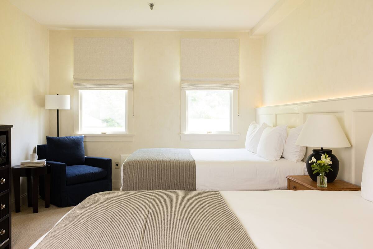A hotel room with two beds, a chair, a nightstand, and a lamp.