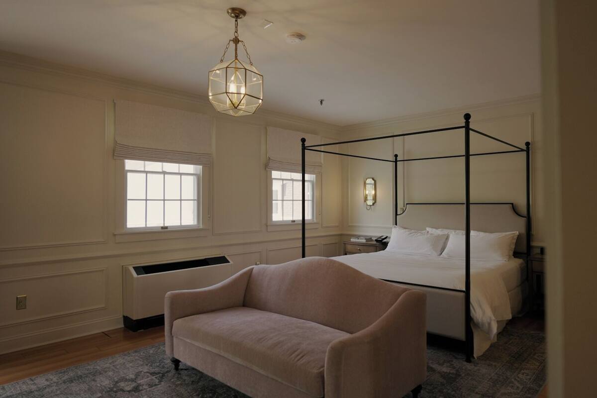 A four-poster bed with white linens, a beige armchair, and a geometric pendant light.