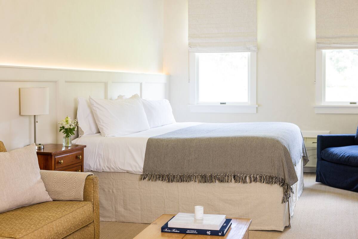 A neatly arranged bedroom with a large bed, a bedside table with a lamp, a small table with a book, and a chair.