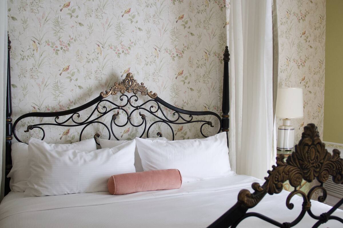 A neatly made bed with white linens and a single pink pillow, a black ornate headboard, and floral wallpaper.