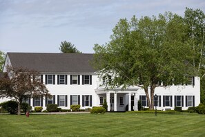 A large, two-story white building with a symmetrical facade, featuring a central entrance with columns, surrounded by well-maintained lawns and mature trees.