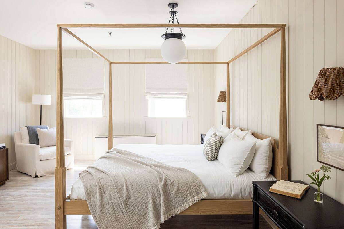 A bedroom with a four-poster bed, a bedside table with a book and a vase, and a chair.