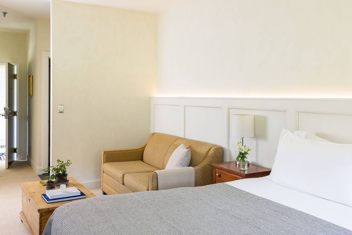 A hotel room with a bed, a sofa, a nightstand with a lamp, and a small table with a book and a plant.