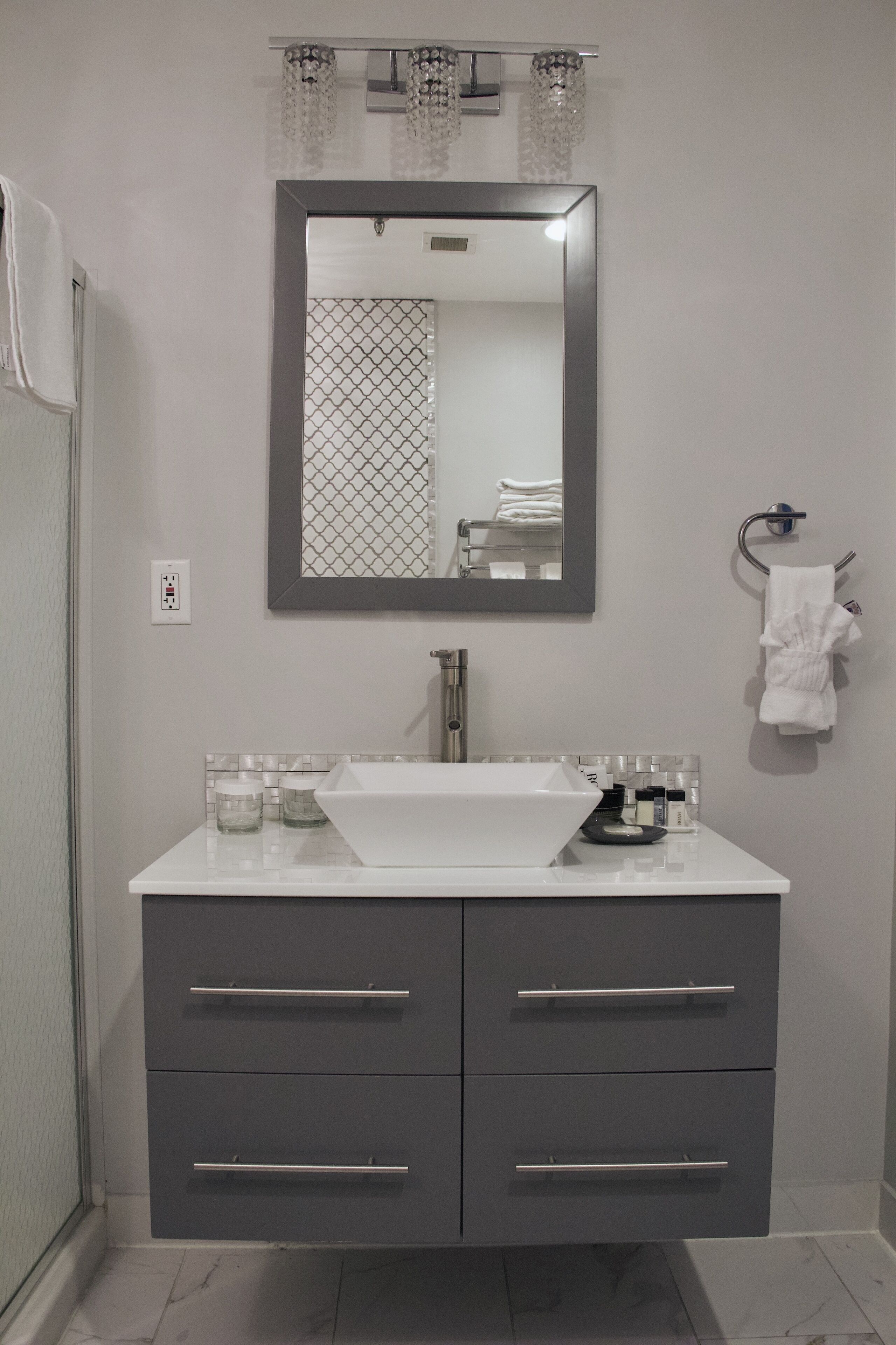 Un baño con un lavamanos blanco, muebles grises, un espejo y un perchero para toallas.