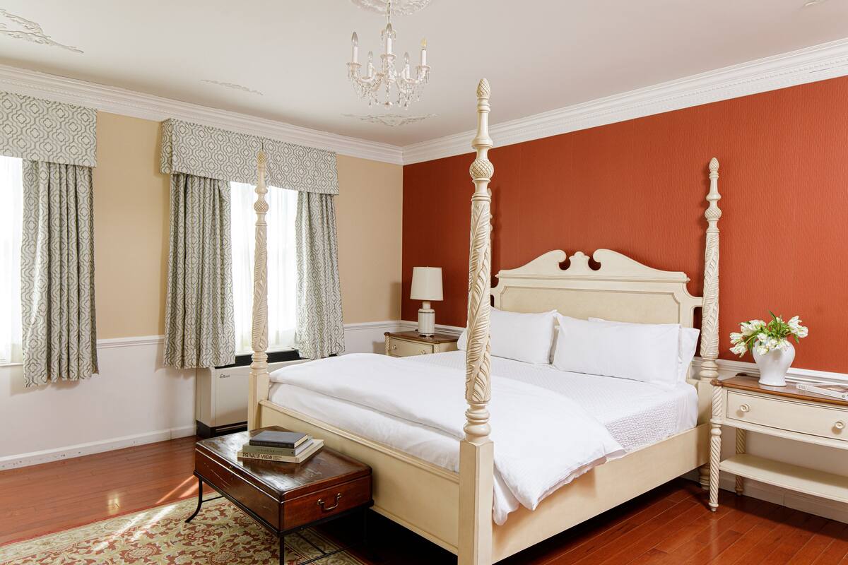 A four-poster bed with a white finish, a nightstand with a lamp, and a bedside table with a book and a vase of flowers.