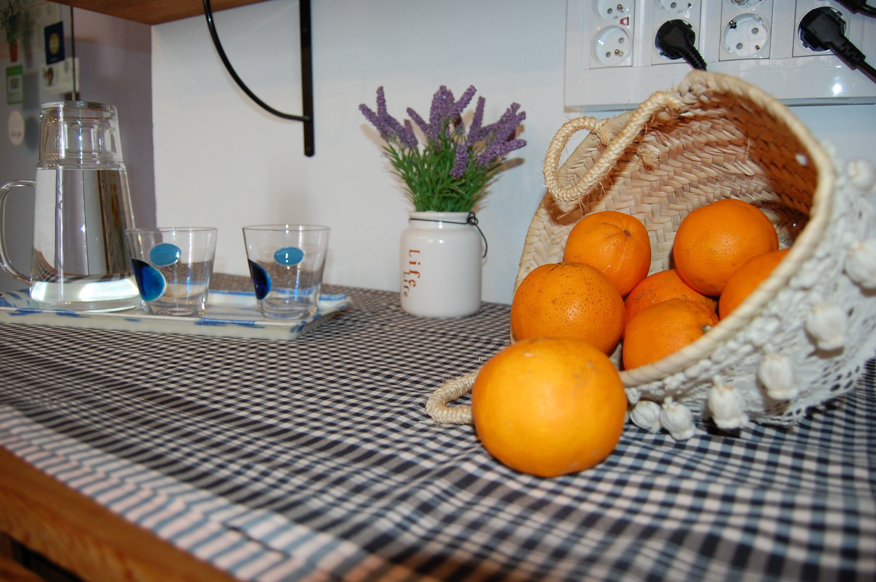 Una encimera de cocina con una jarra, vasos, un florero con lavanda y una cesta de naranjas.