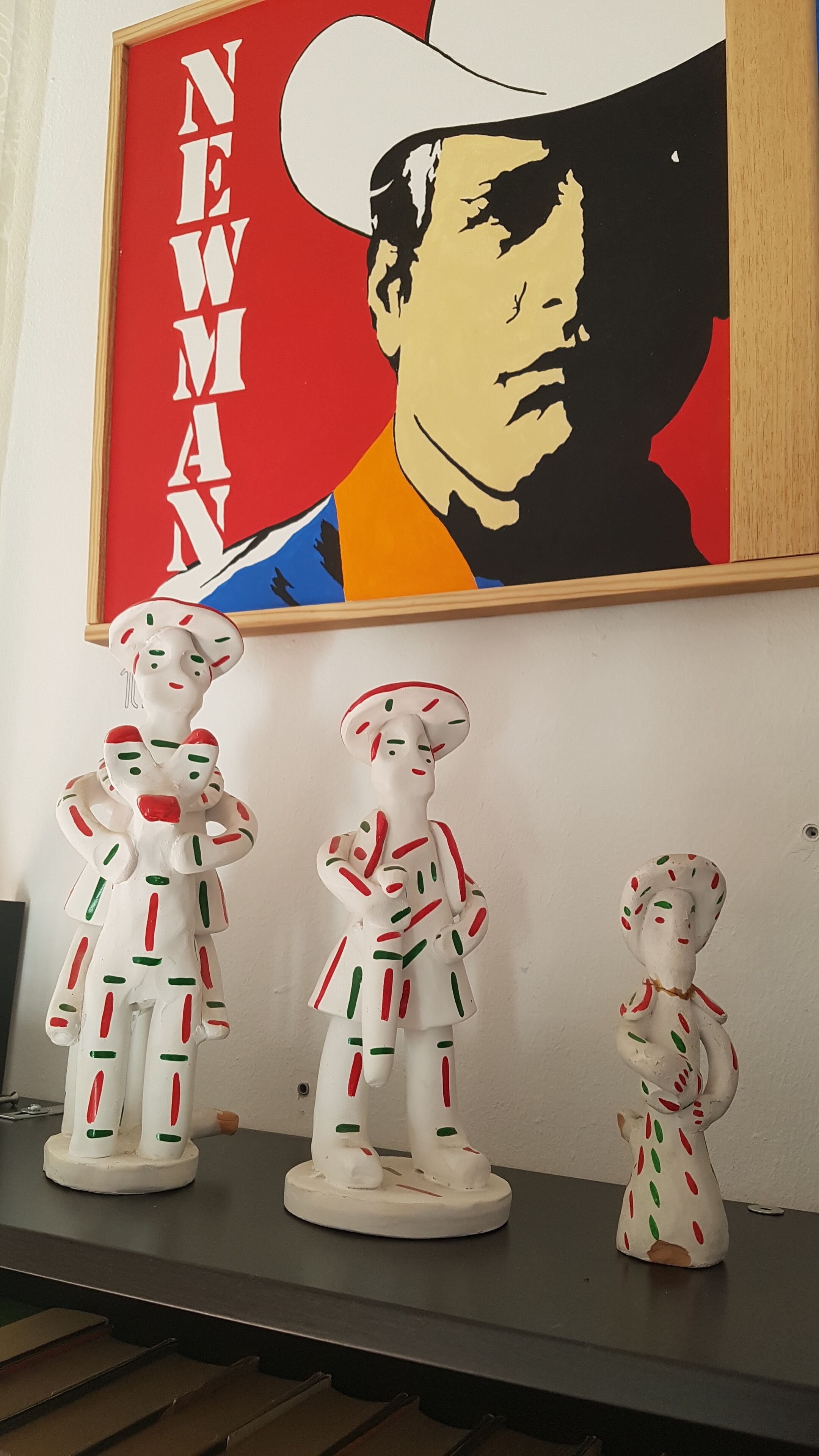 Una obra de arte enmarco con la palabra "NEWMAN" y un retrato de un hombre. Abajo, hay tres figuritas de cerámica con rayas rojas y verdes.