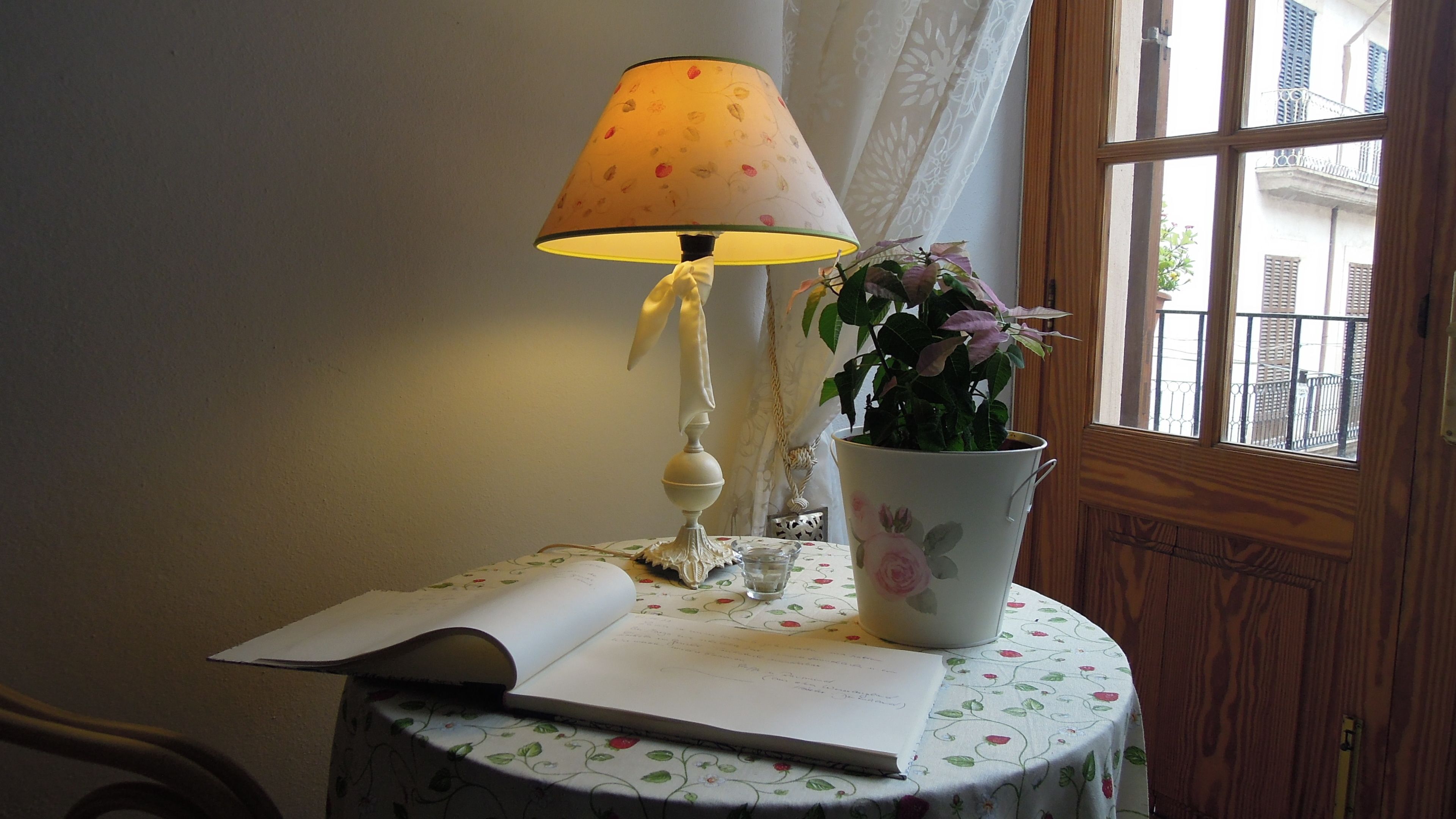 A table with a floral tablecloth, a rolled-up book, a white lamp with a floral shade, and a potted plant.