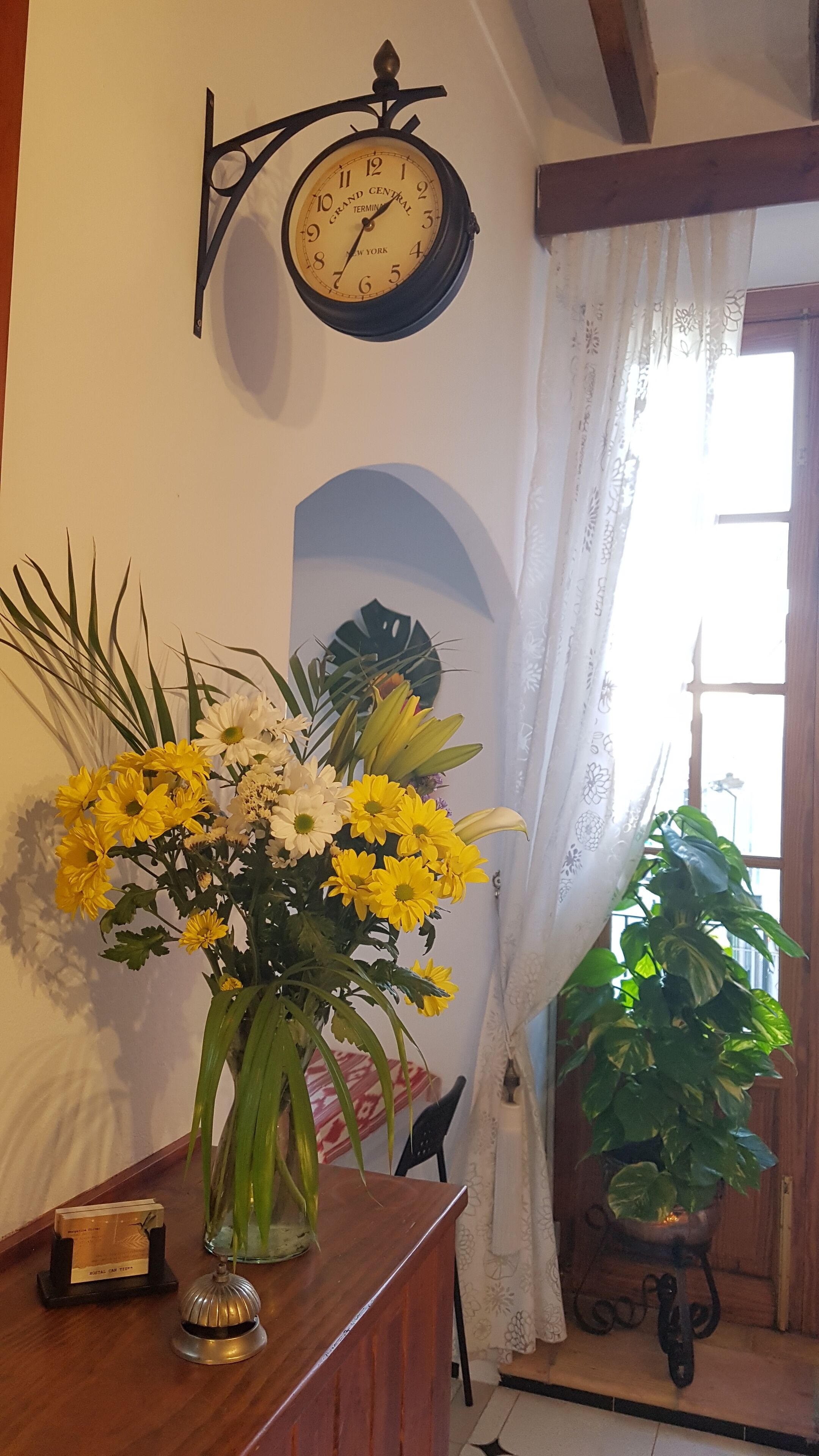 Una habitación con un escritorio de madera, un reloj, un arreglo floral y una planta en maceta.