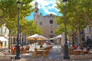 An outdoor café with tables and chairs, surrounded by trees and a historic building with a clock tower in the background. - Hostal Ca'n Tiu (Alaro)