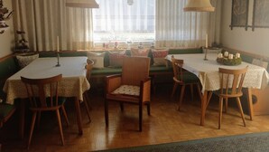 Un comedor con un amplio ventanal, un banco acolchado verde, sillas de madera y mesas cubiertas con manteles blancos.