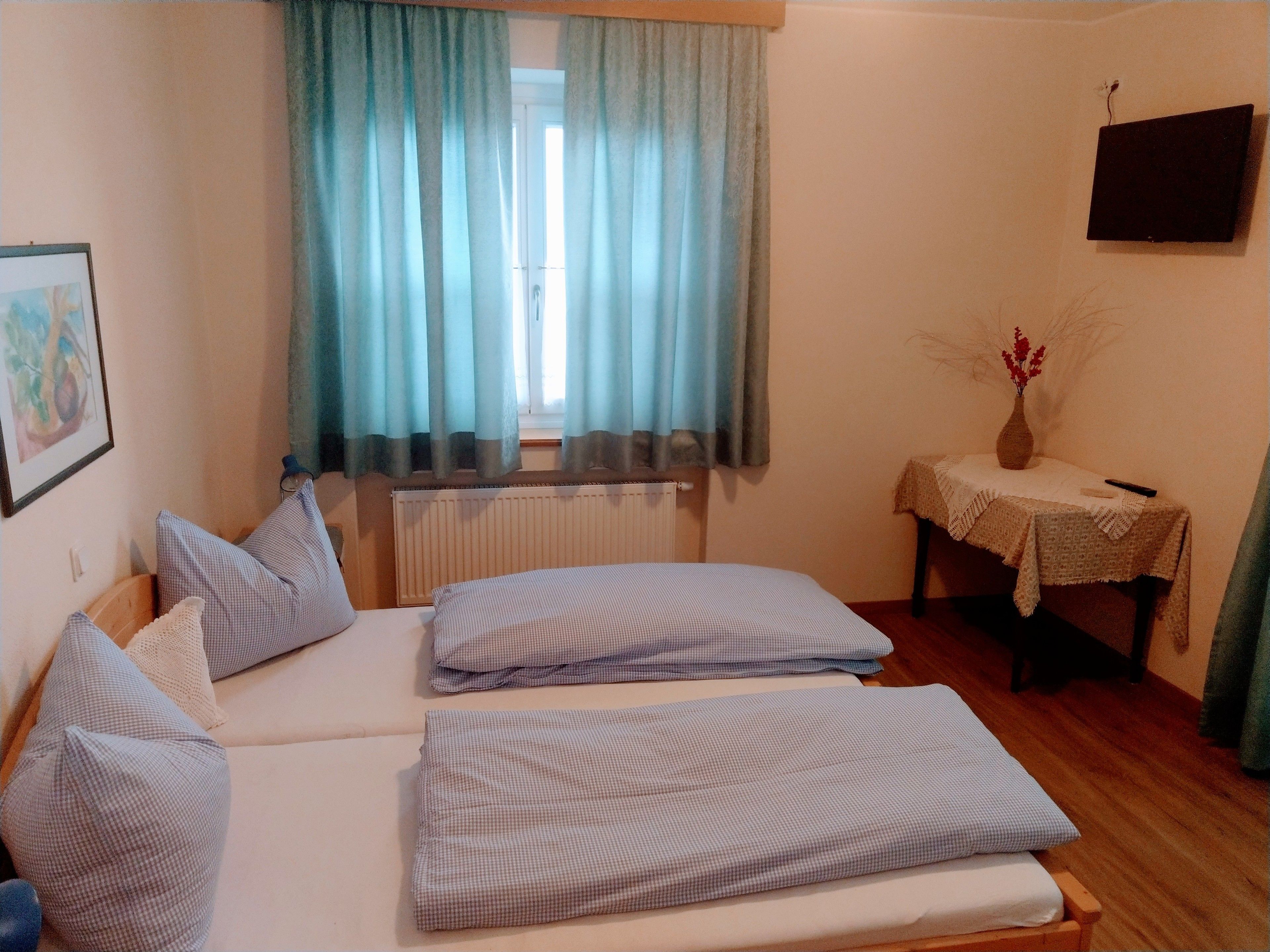 Um quarto com cama, almofadas, radiador, mesa com um vaso e um quadro na parede.