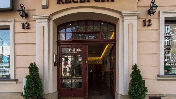L’entrée d’un immeuble, au-dessus de la porte, on peut lire l’inscription « RECEPCJA ». De part et d’autre de l’entrée, on trouve deux plantes en pot.