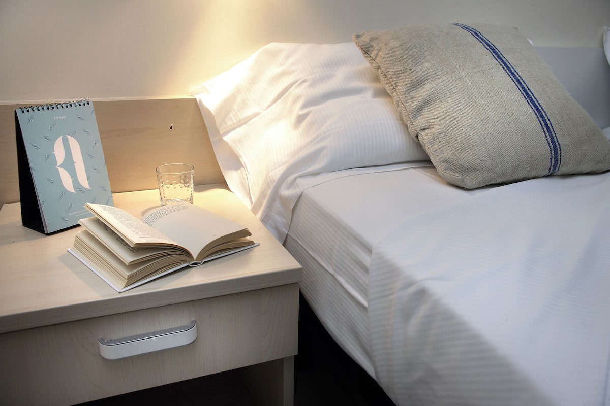 A neatly made bed with white linens, a pillow with a blue stripe, a bedside table with a book, a glass of water, and a calendar.