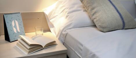 Cama bem arrumada com lençóis brancos, travesseiro com faixa azul, mesa de cabeceira com um livro, um copo d'água e um calendário.