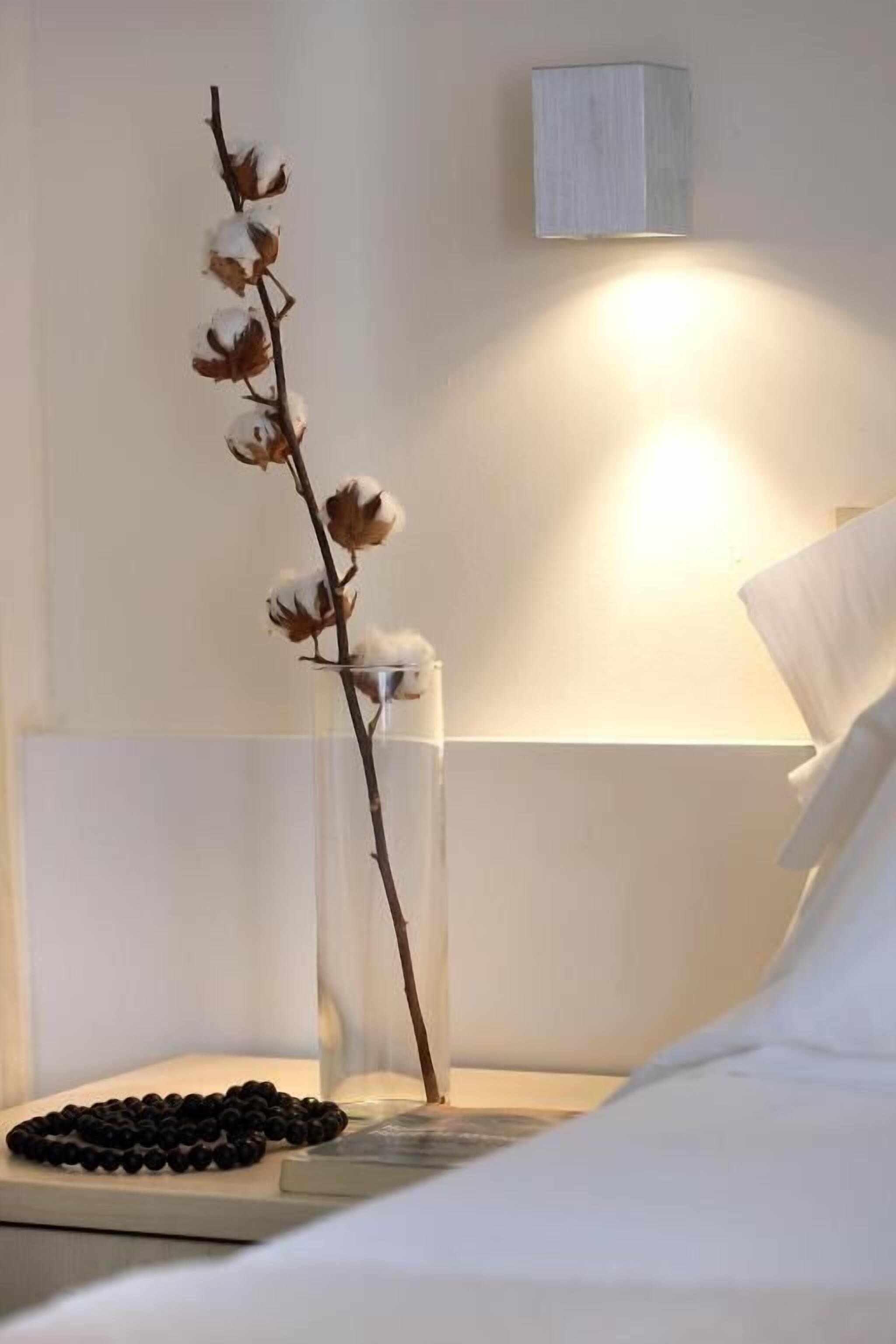 A neatly made bed with white linens, a bedside table with a clear vase containing a branch with cotton flowers, and a wall-mounted light fixture.