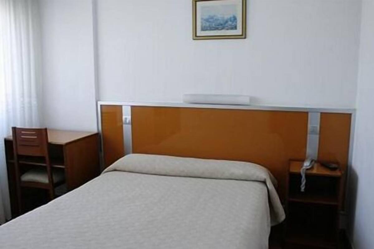 Quarto de hotel com uma cama de solteiro, uma escrivaninha de madeira, uma cadeira e uma pequena mesa de cabeceira.