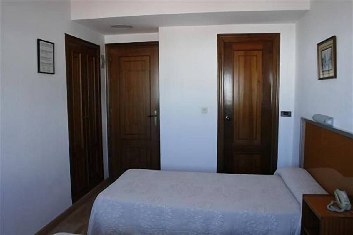 Quarto com uma cama, duas portas de madeira, uma mesa de cabeceira e uma pequena escrivaninha com um telefone.