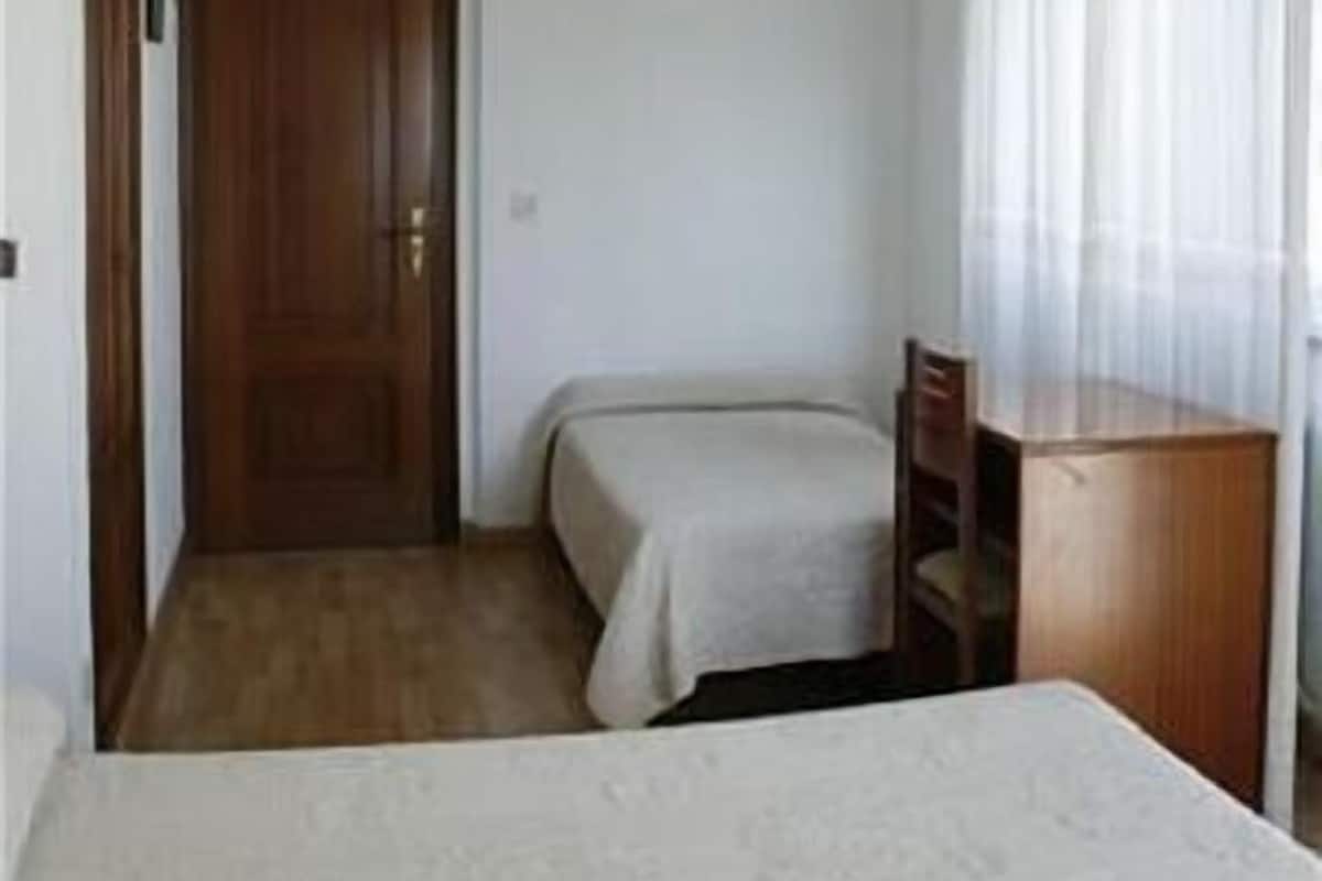 Quarto de hotel pequeno e individual, com uma cama, uma escrivaninha e uma cadeira.