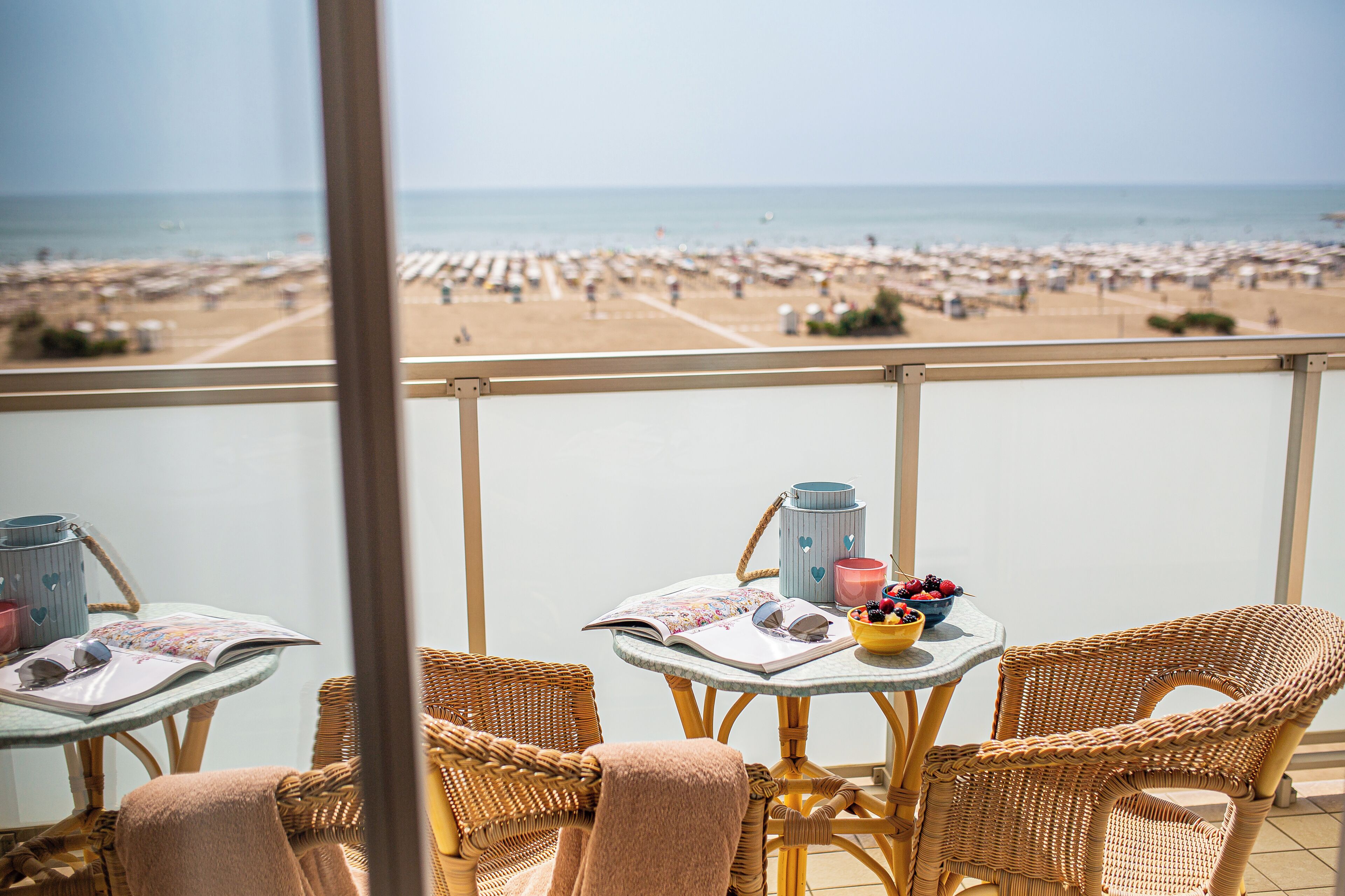 Un balcon avec des fauteuils en osier et une table, donnant sur une plage parsemée de parasols et fréquentée par de nombreuses personnes.