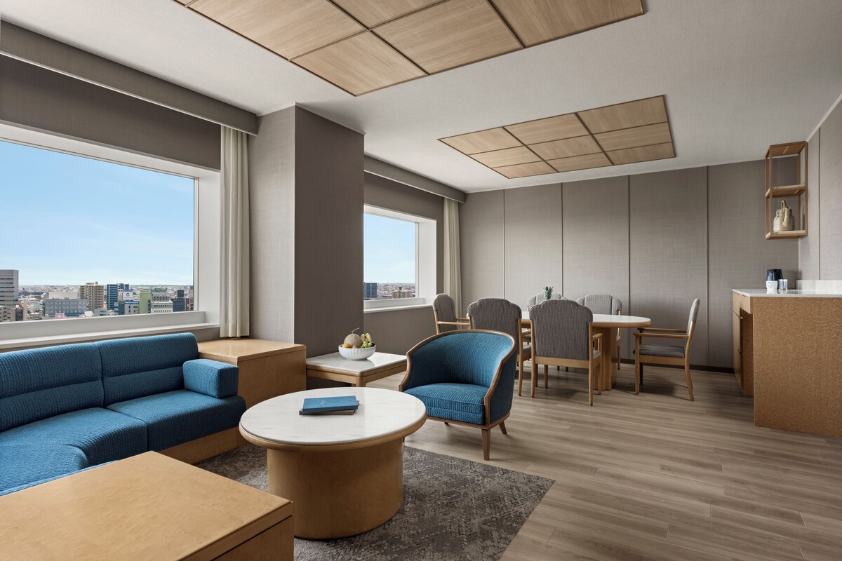 A modern hotel room with a sofa, chairs, a dining table, and a view of the city.