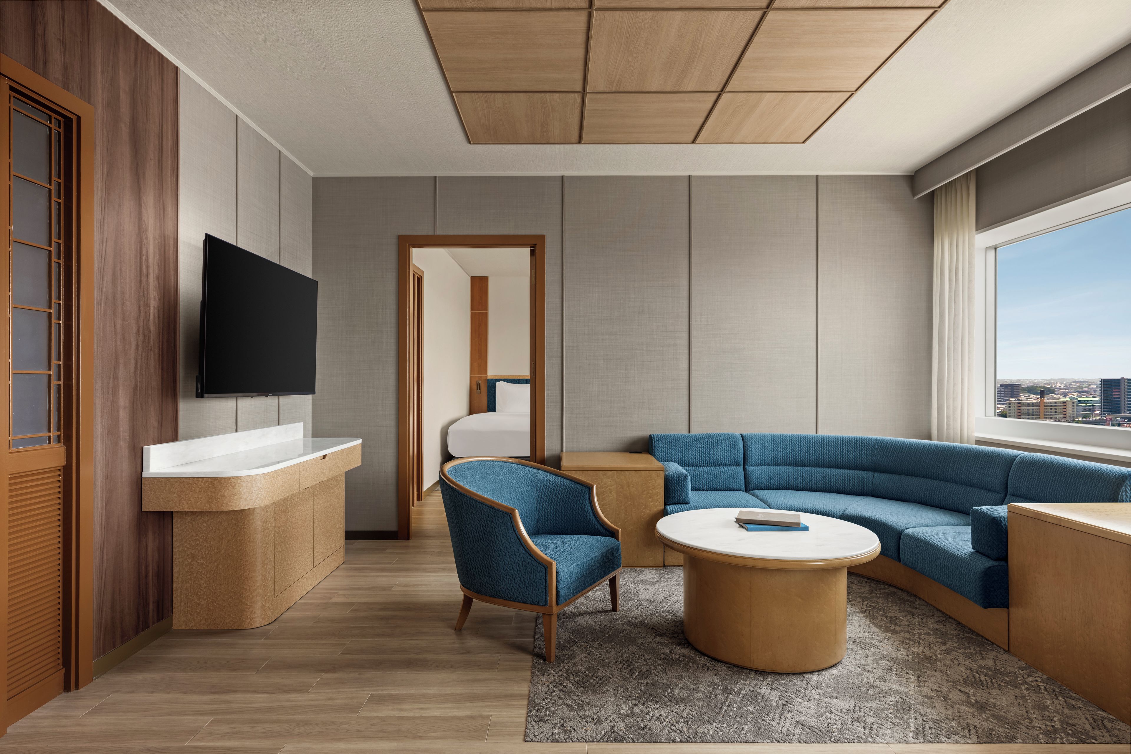 A modern hotel room with a wooden floor, a flat-screen TV mounted on the wall, a sofa, a coffee table, and a view of the city through a large window.
