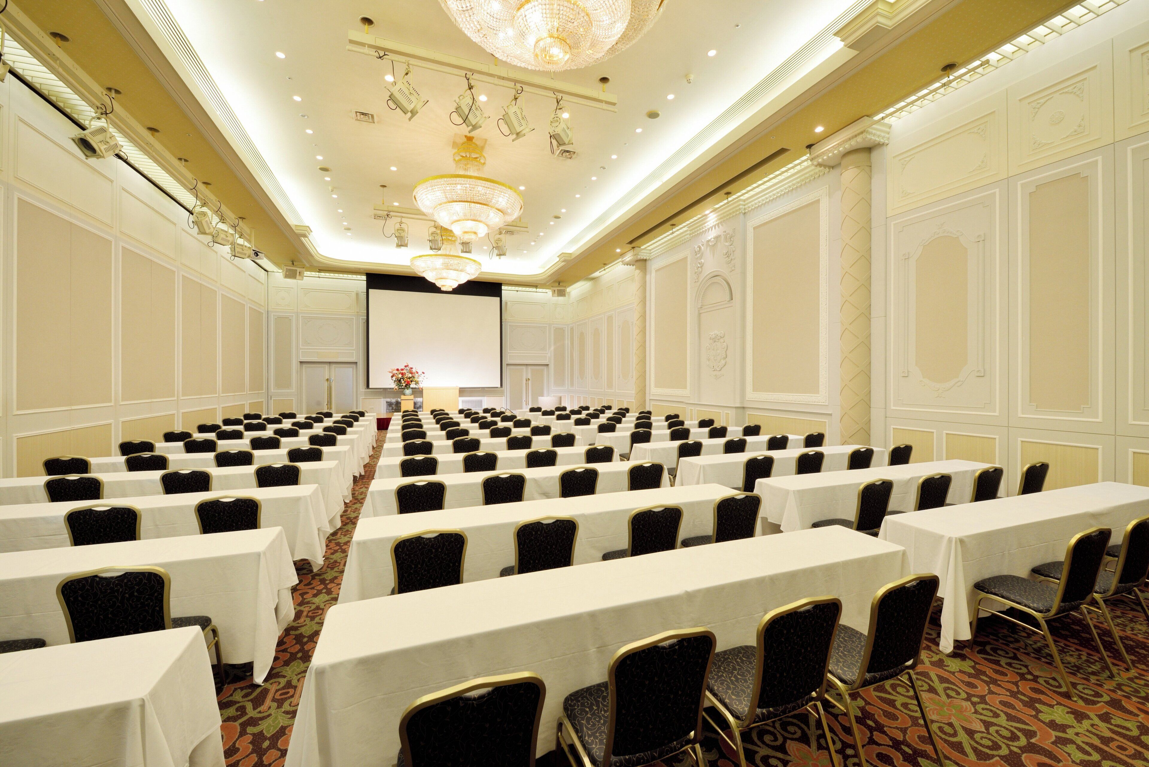 Un salón de conferencias con filas de mesas y sillas, una pantalla grande al frente y detalles ornamentales en el techo.