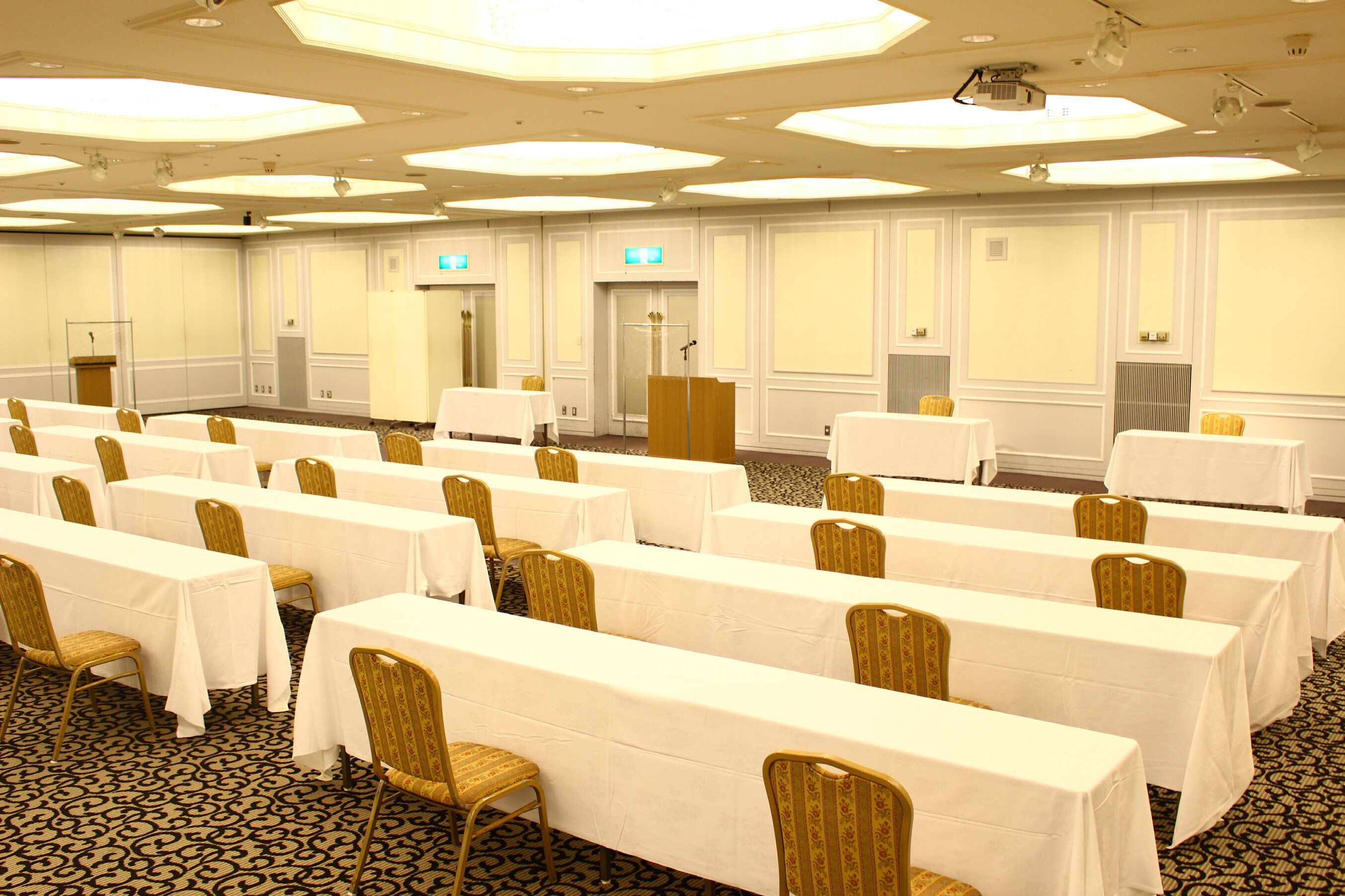 A conference room with rows of tables and chairs, a podium, and a carpeted floor.