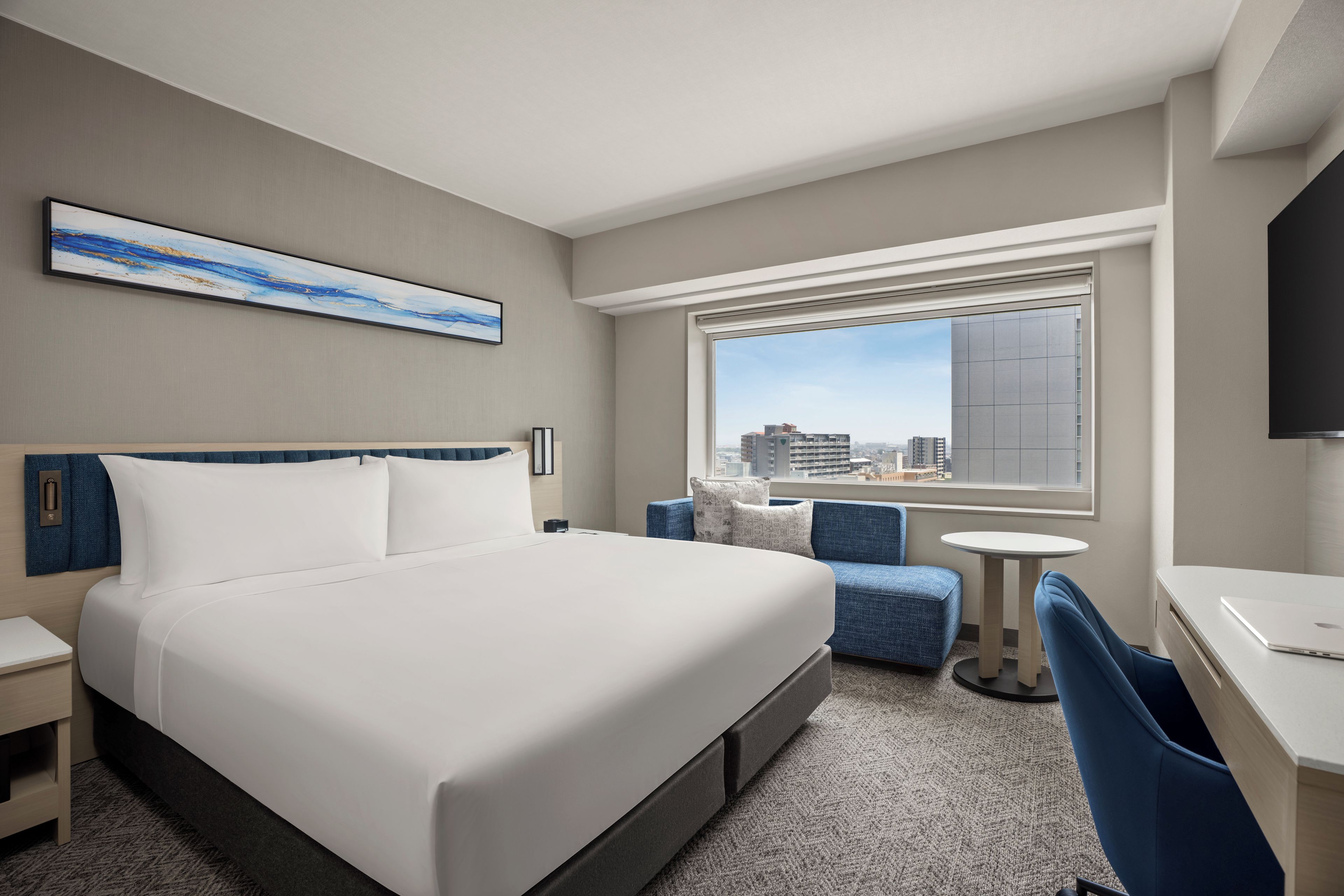A hotel room with a bed, a desk, a chair, a small table, a television, and a window with a city view.