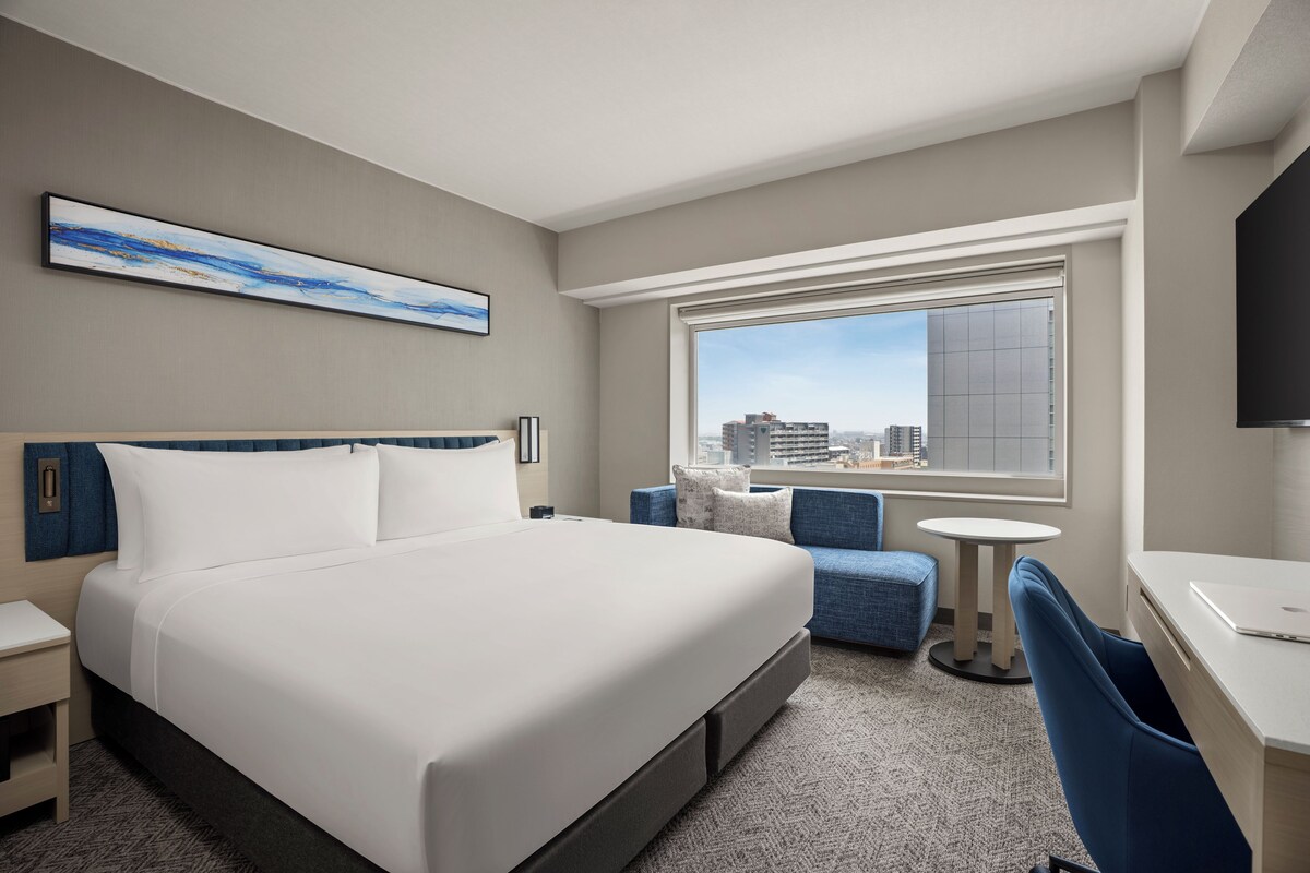 A hotel room with a bed, a desk, a chair, a small table, a television, and a window with a city view.