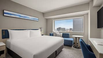 A hotel room with a bed, a desk, a chair, a small table, a television, and a window with a city view.