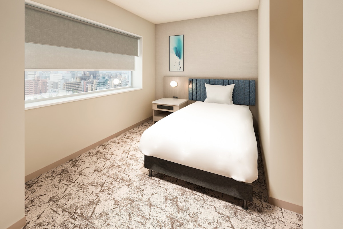 A hotel room with a bed, a nightstand, a window with a city view, and a painting on the wall.