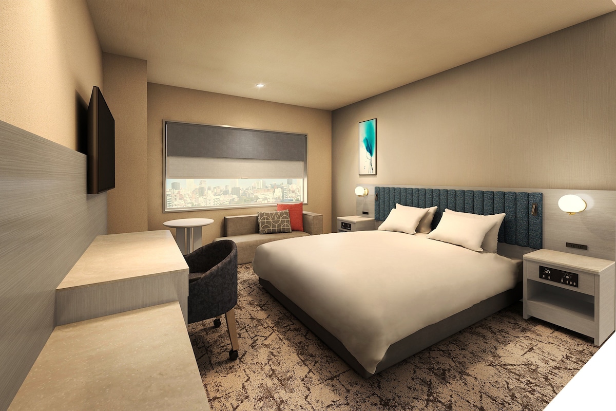 A modern hotel room with a large bed, a desk, a chair, a nightstand, and a window with a city view.
