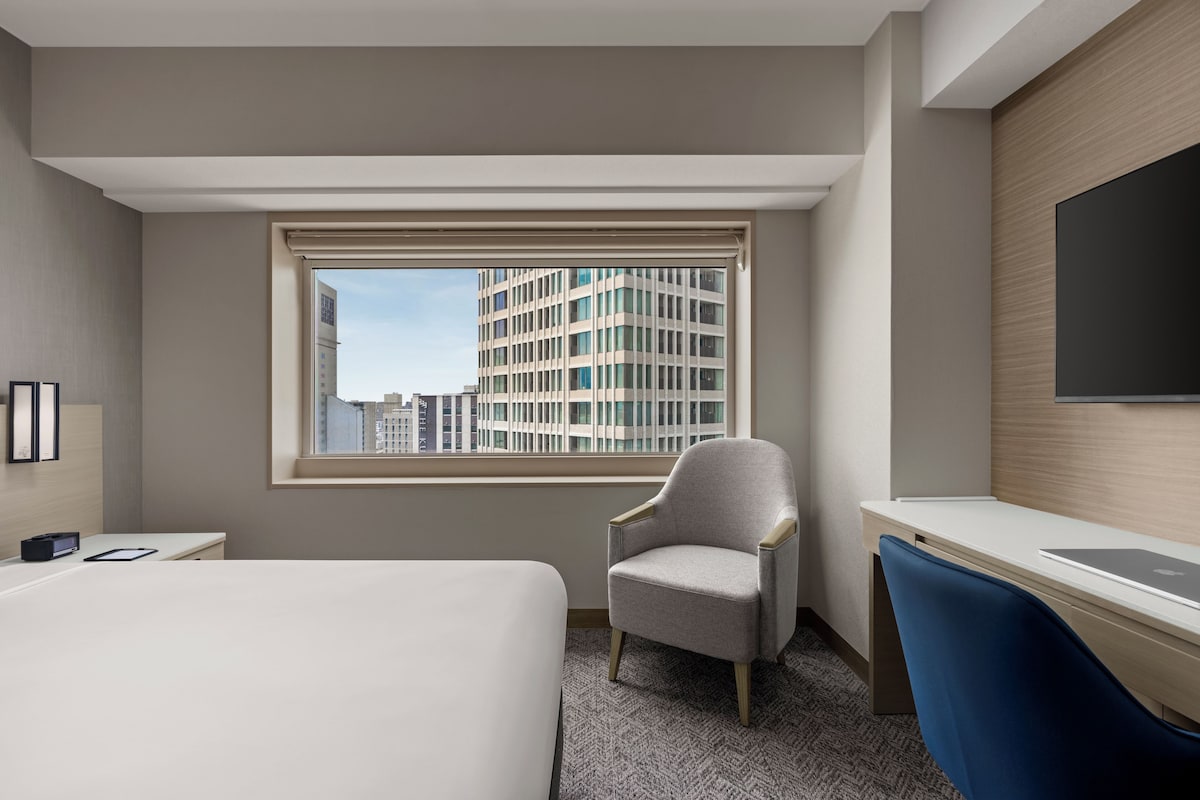 Quarto de hotel com cama, escrivaninha, cadeira, janela grande com vista para a cidade e televisão de tela plana fixada na parede.