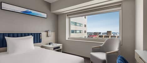 Habitación de hotel con cama, escritorio, silla, mesita de noche, ventana con vistas a la ciudad y un cuadro en la pared.