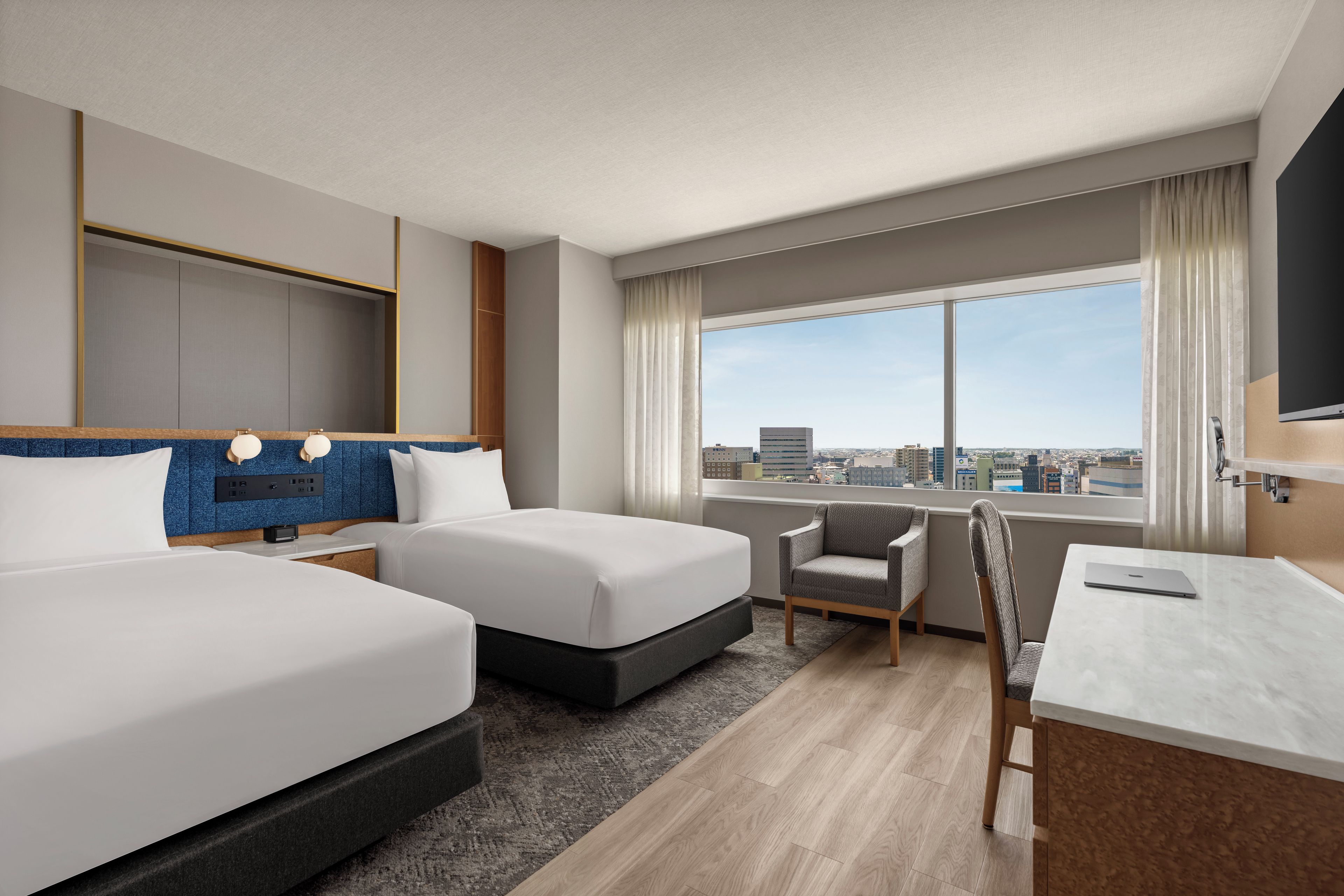 A hotel room with two beds, a desk, a chair, and a large window with a city view.