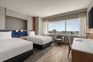 Ein Hotelzimmer mit zwei Betten, einem Schreibtisch, einem Stuhl und einem großen Fenster mit Blick auf die Stadt.
