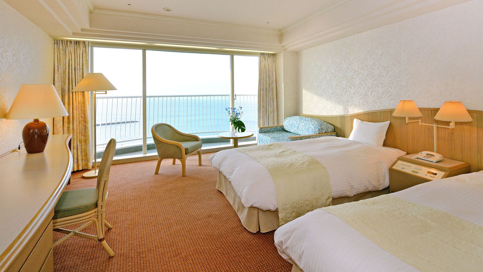 Une chambre d’hôtel avec deux lits, une chaise, un bureau, une table de chevet et une vue sur la mer grâce à de grandes fenêtres.