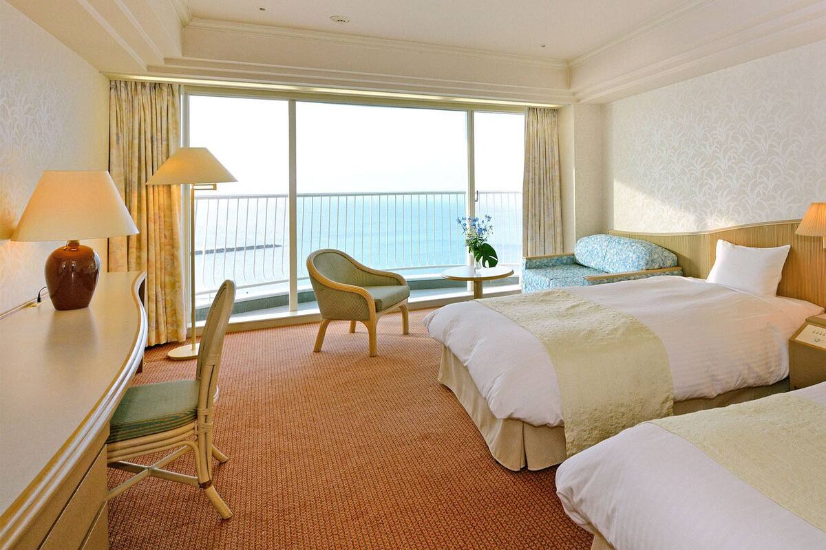A hotel room with two beds, a chair, a desk, a nightstand, and a view of the sea through large windows.