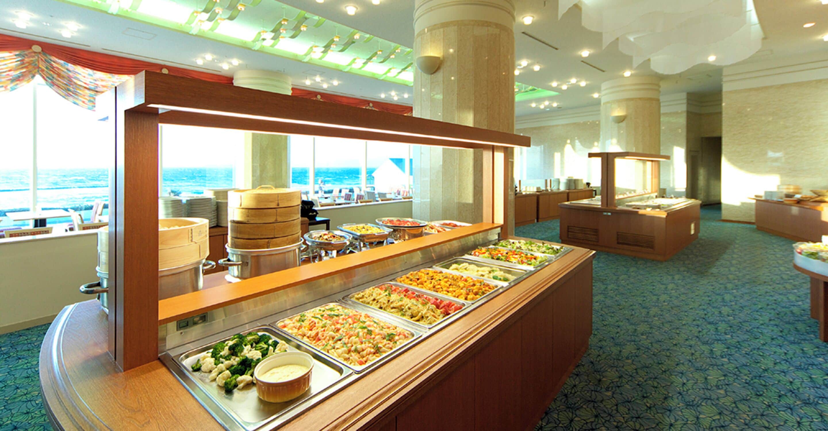 A buffet area with multiple food stations, a view of the ocean through large windows, and a patterned carpet.