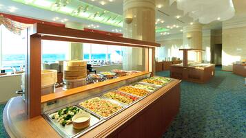 A buffet area with multiple food stations, a view of the ocean through large windows, and a patterned carpet.