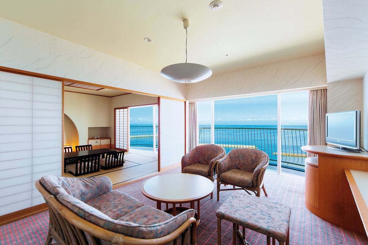 A spacious room with a large window offering a view of the sea, a round table, two armchairs, a sofa, and a television.