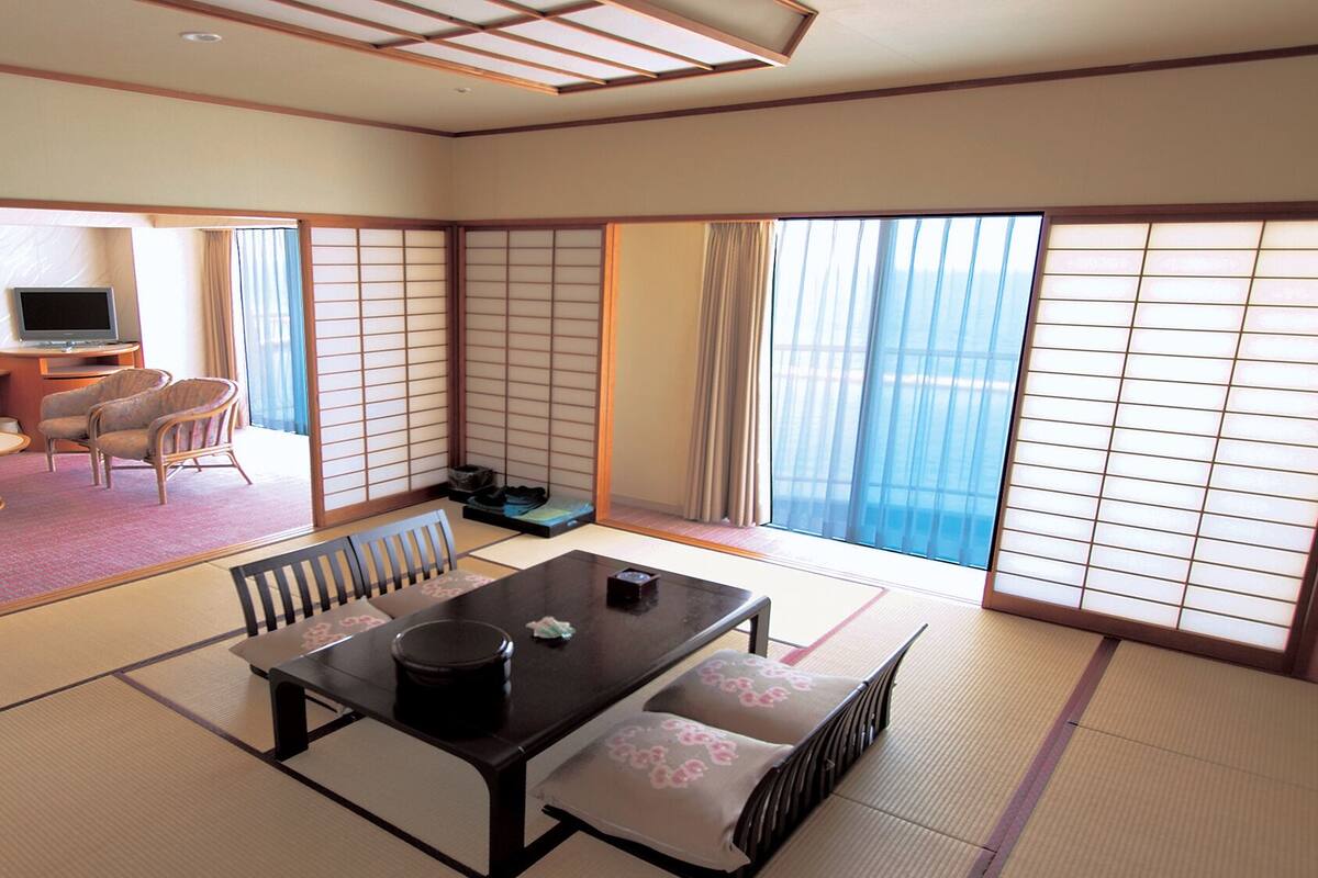 A traditional Japanese room with tatami flooring, a low table with cushions, a television, and a view of the sea.