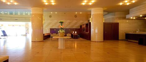 A spacious lobby with a central reception desk, a large vase of flowers, and a modern design with tiled flooring and ceiling lights.