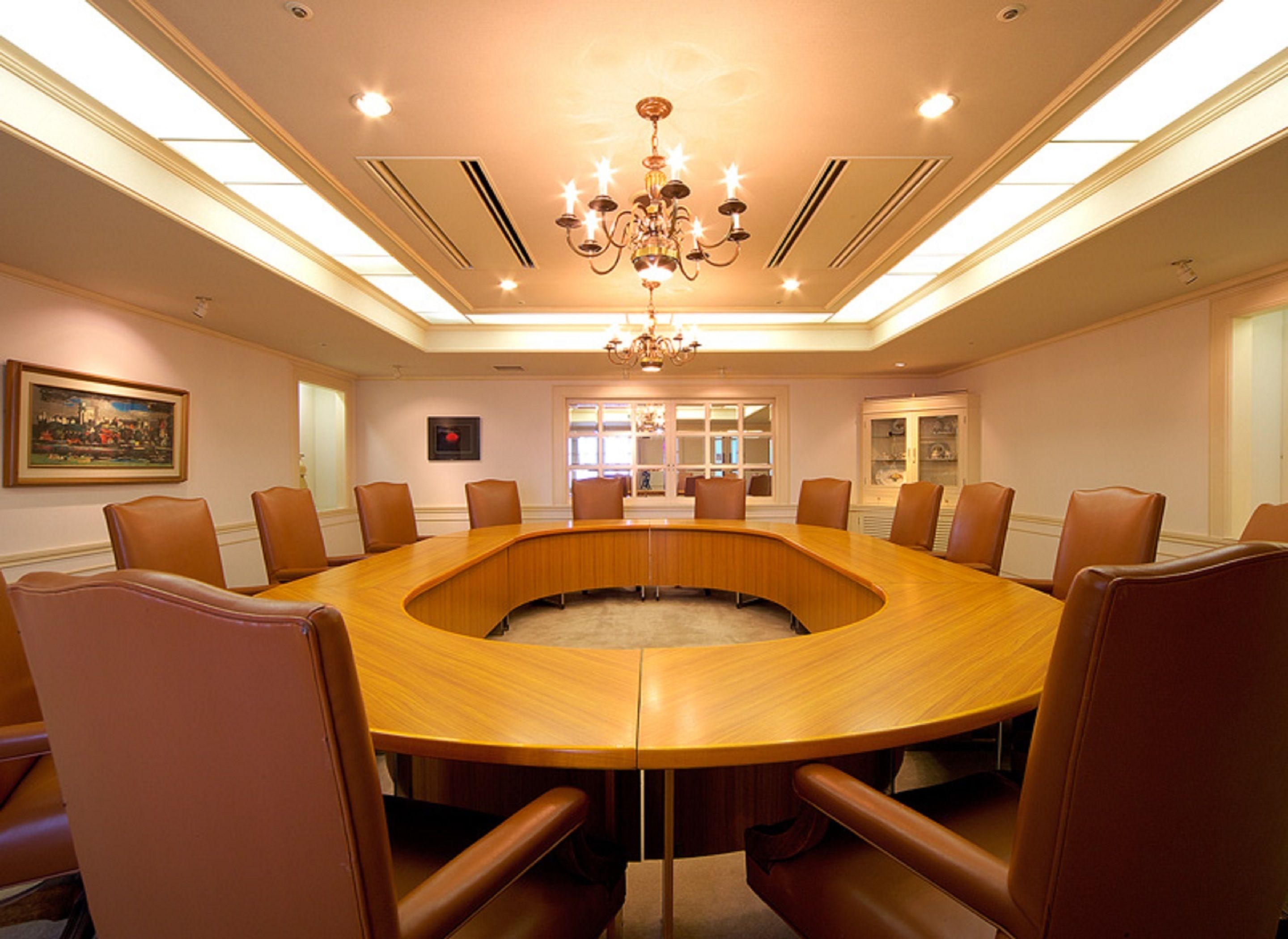A conference room with a round table, leather chairs, a chandelier, and a painting on the wall.